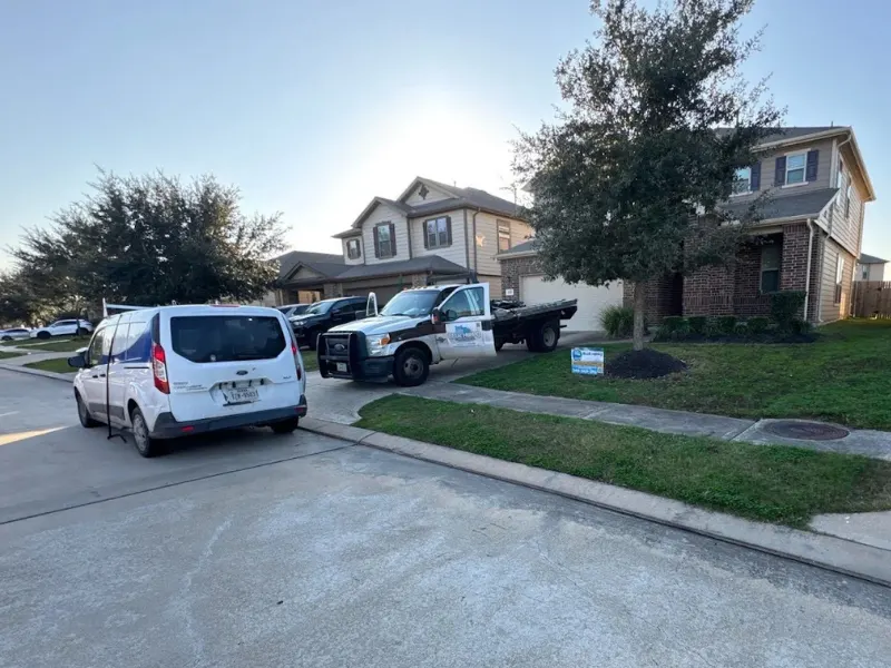 A white van and a flatbed truck parked in front of suburban houses with a tree on the lawn.