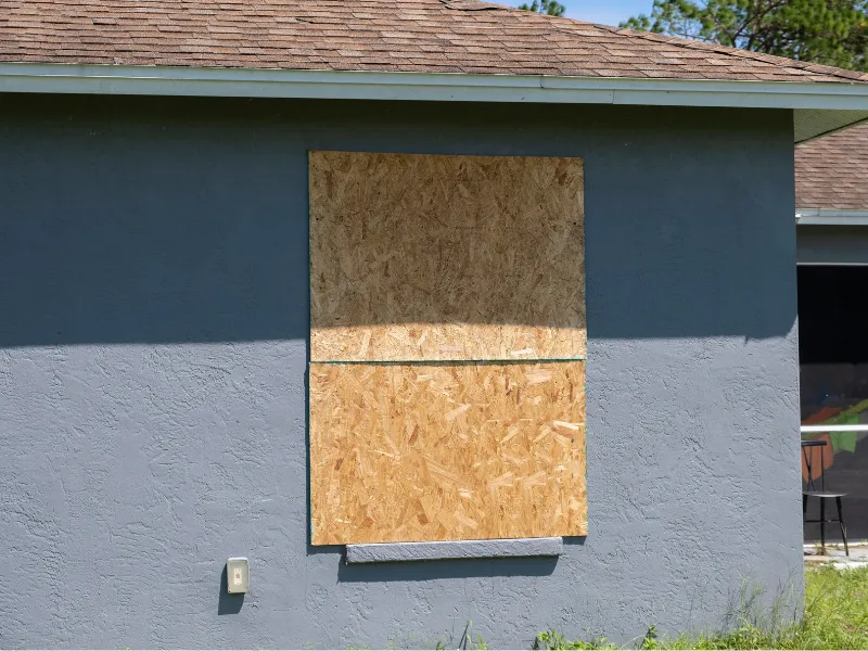 Window boarded up with plywood on a blue-gray house exterior.