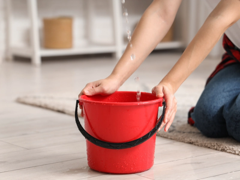 Hands holding a red bucket on the floor as water is poured into it.