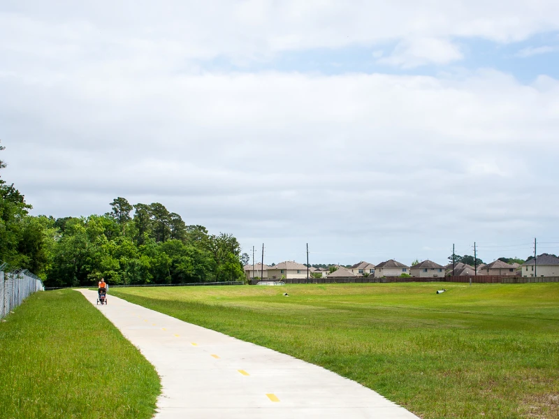 Person walking with a stroller on a paved path beside a grassy field and houses in the distance.