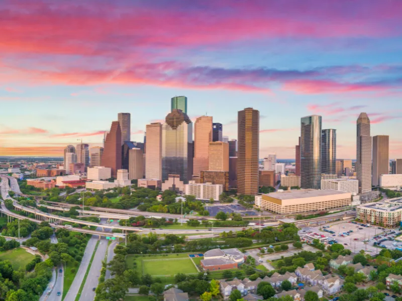 Houston skyline with highways and green spaces under a colorful sunset sky.