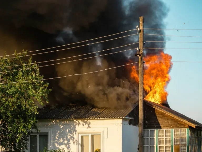 Flames and thick black smoke rising from the roof of a burning house near a utility pole.