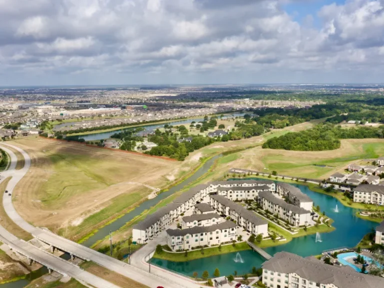Aerial view of a residential complex surrounded by water canals and green fields under a cloudy sky.