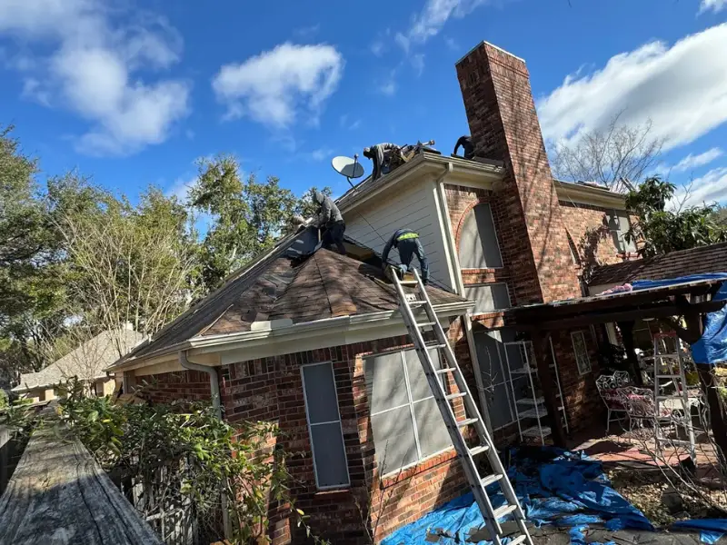 Three workers repair a shingled roof on a brick house under a blue sky.